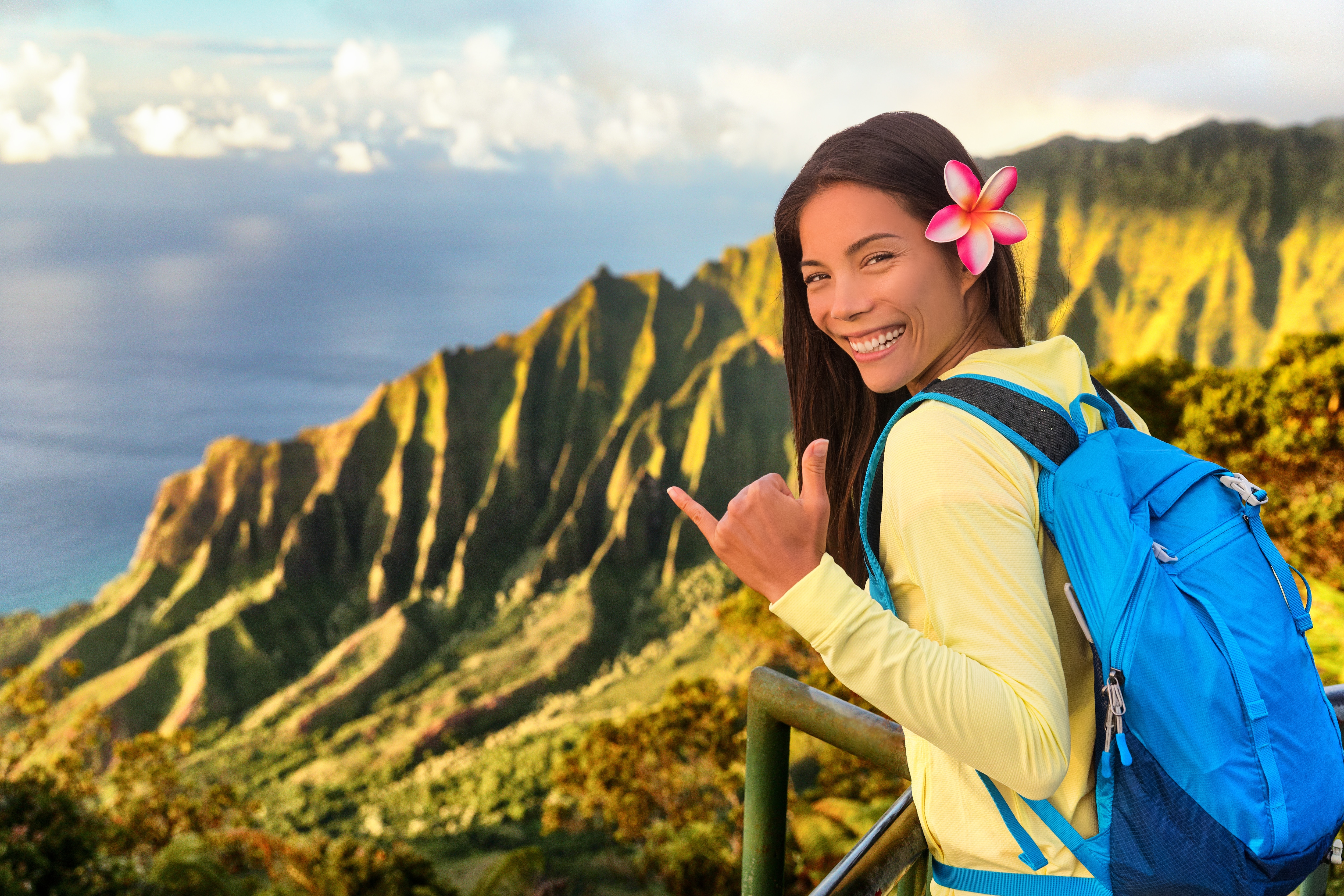 Hawaii hiker girl tourist doing shaka hawaiian hand sign at Na pali lookout in Kauai, Hawaii. Travel Asian woman backpacker hiking with bag at famous destination Kalalau hawaiian attraction.