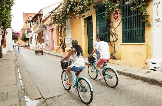 2 people riding colorful bikes on a small street next to colorful houses covered with flowers cascading from the rooftops