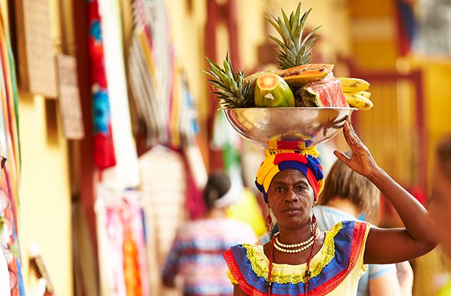 A woman dressed in colorful, native attire balancing a bowl of fruit on her head