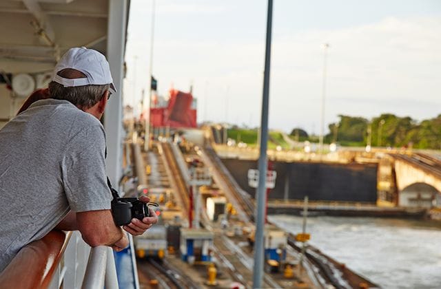 Man leaning over the railing of a ship, holding his camera, viewing the locks of the Panama Canal
