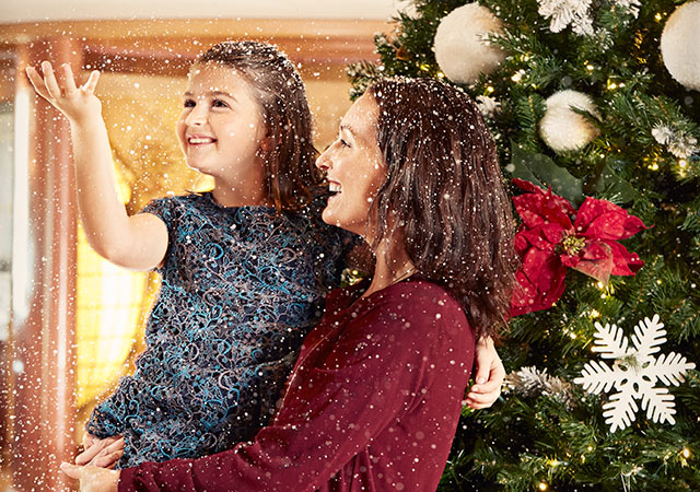 mother holds daughter in front of a decorated Christmas tree. Daughter throws artificial snow in the air.